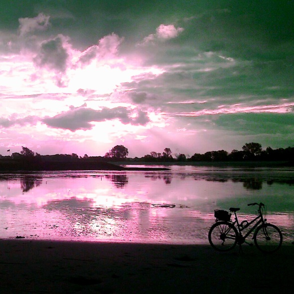 sonnendurchfluteter wolkenhimmel in pink, darunter wasser, ein fahrrad davor auf dem strand